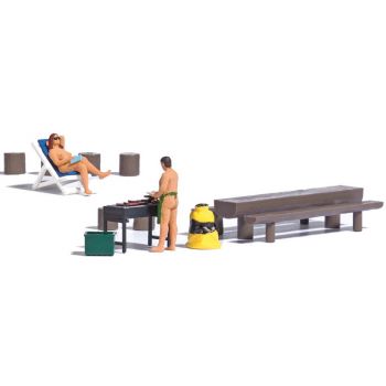 Nacktgriller - Nackter Mann am Grill nur mit einer Schürze bekleidet und nackte Frau im Liegestuhl, mit Grill und Grillgut und div. Zubehör