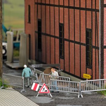 Absperrungen aufstellen, 3 Straßenbauarbeiter in Arbeitskleidung mit verschiendenen Absperrungen, Schildern, Schubkarren, Werkzeug und Sandhaufen,