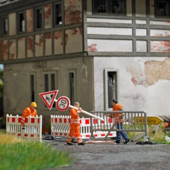Absperrungen aufstellen, 3 Straßenbauarbeiter in Arbeitskleidung mit verschiedenen Absperrungen, passenden Verkehrsschildern, Pylonen, Sandhaufen usw.